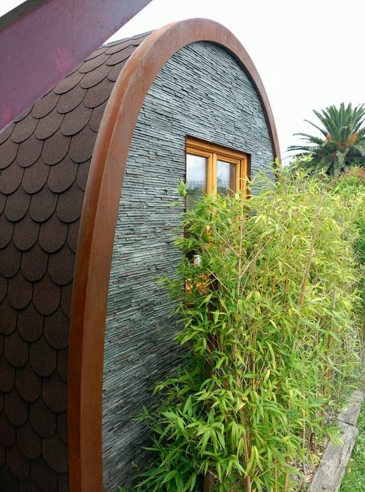 Fachada de bungalow prefabricado Maxicozy con piedra pizarra, acero Corten y tejado curvo de escamas